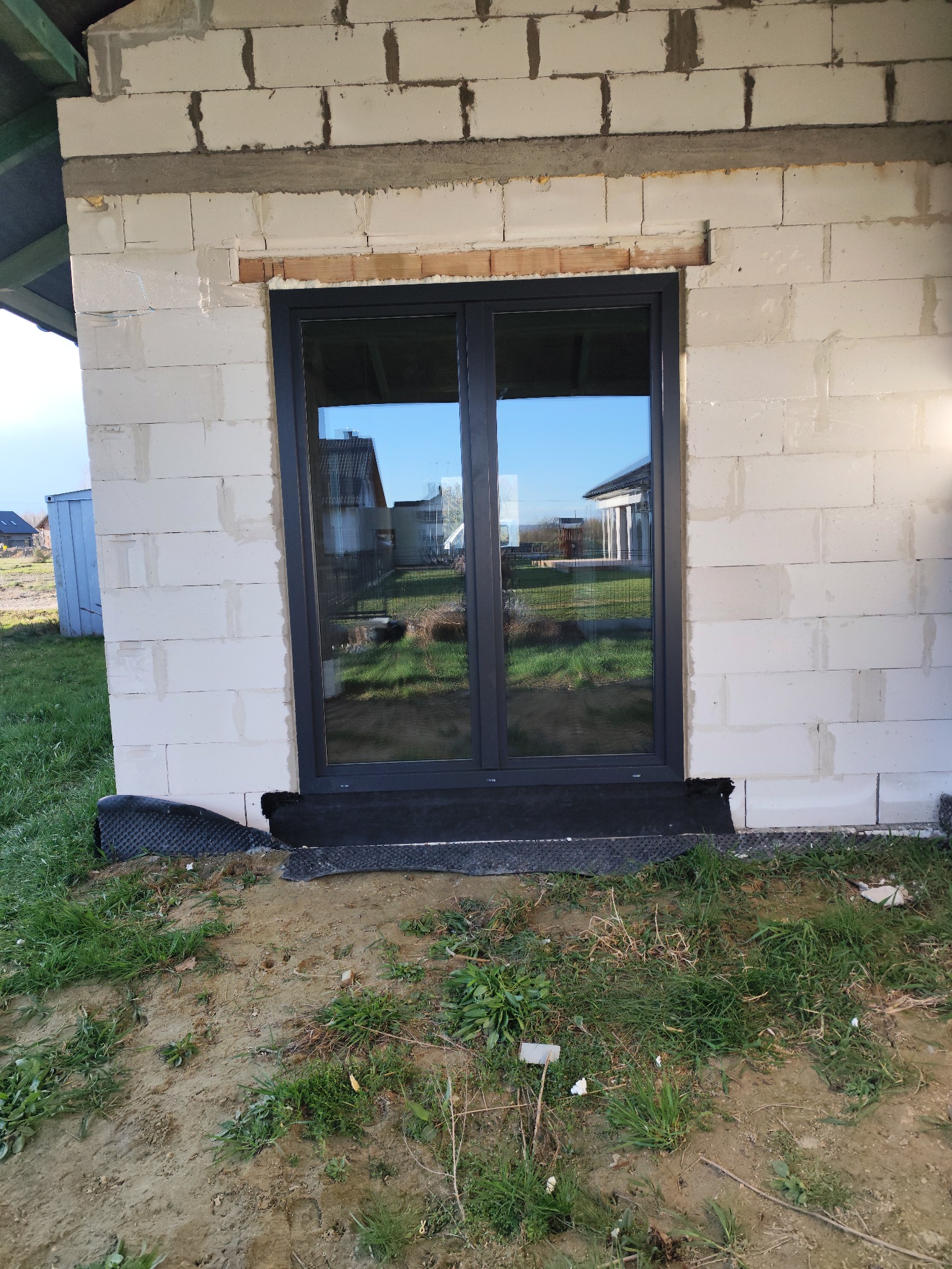 Zamontowane, grafitowe okno PCV w surowym murze z betonu komórkowego. W szybach odbija się krajobraz. Widoczne czarne uszczelnienie wokół ramy.