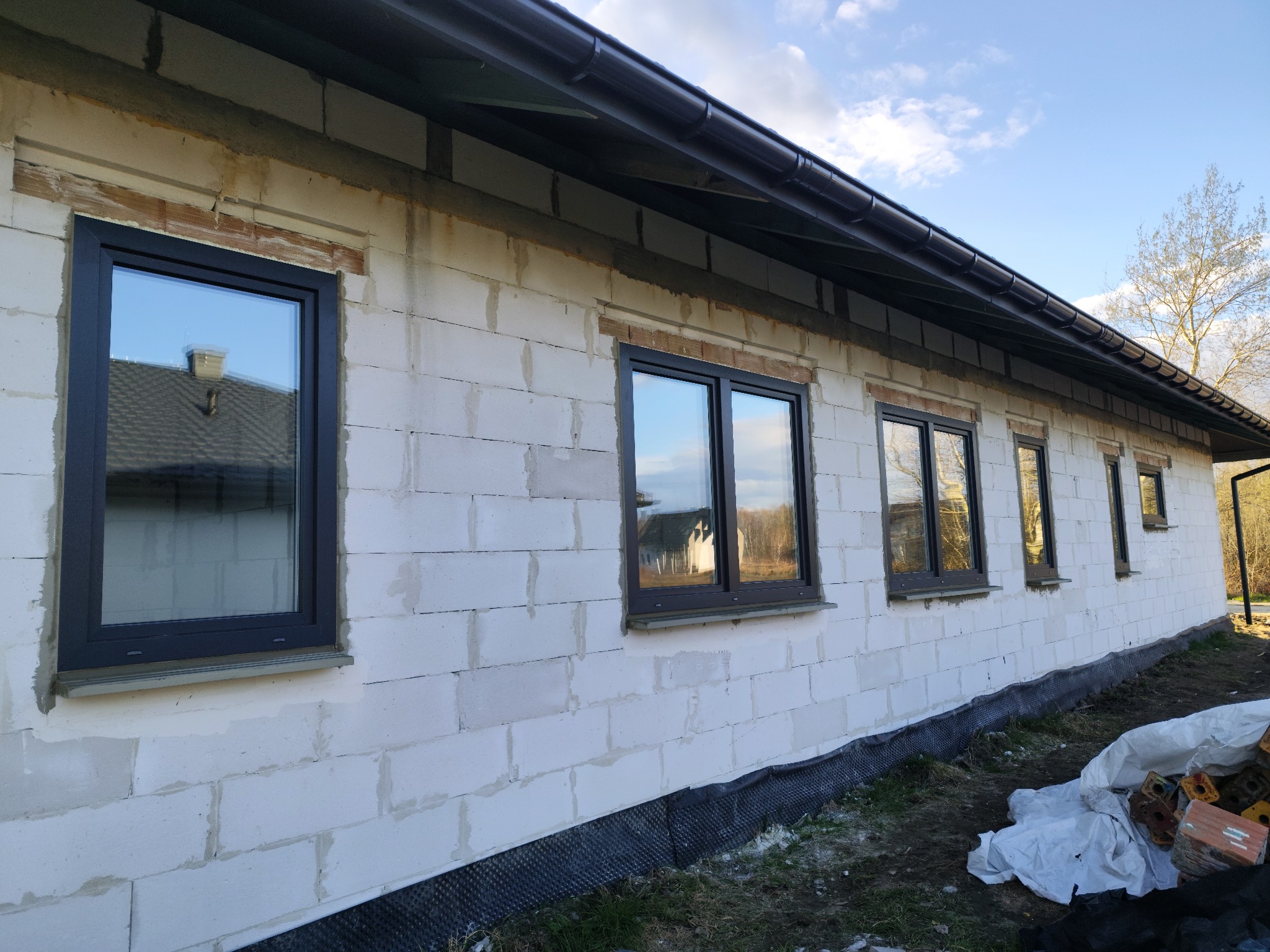 Surowy mur z zamontowanymi oknami w ciemnych ramach, widok z zewnątrz. Okna z parapetami, na dole widoczna folia fundamentowa. Dach z rynnami.