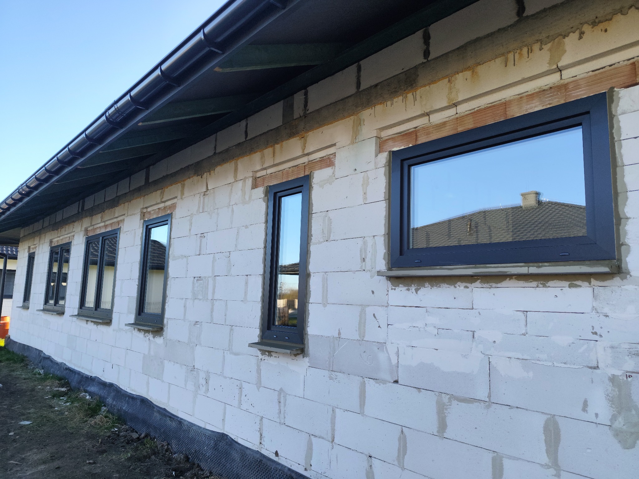 Surowy budynek z zamontowanymi, grafitowymi oknami. Widoczne bloczki z betonu komórkowego i fragment dachu odbijający się w szybach okien.