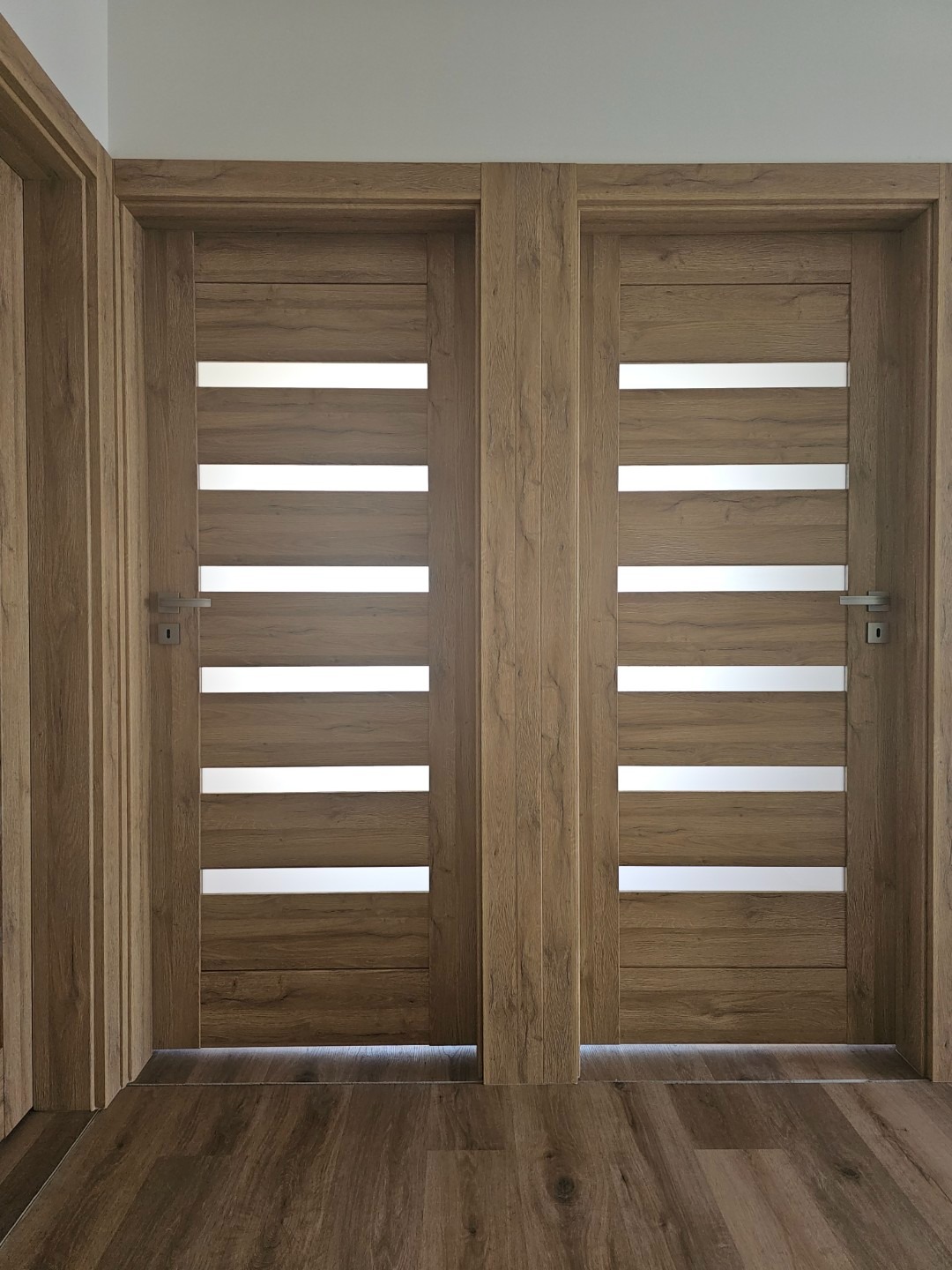 Dwa skrzydła drewnianych drzwi wewnętrznych z poziomymi, mlecznymi przeszkleniami i srebrnymi klamkami, zamontowane w jasnym wnętrzu z podłogą imitującą drewno.