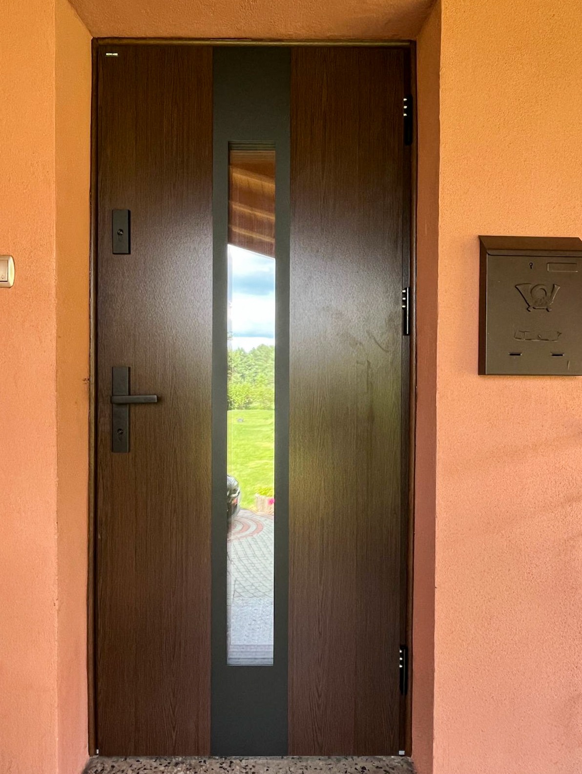 Ciemnobrązowe drzwi wejściowe z wąskim, pionowym przeszkleniem, zamontowane w pomarańczowej ścianie budynku. Widoczna skrzynka pocztowa i fragment podjazdu.