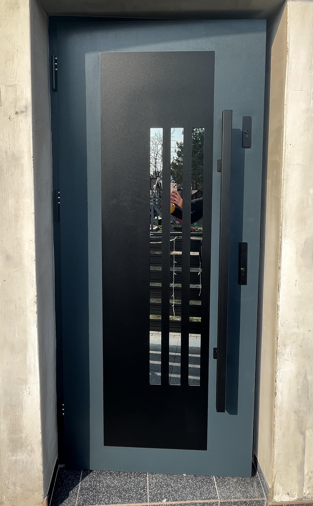 Nowoczesne, grafitowe drzwi zewnętrzne z podłużnym przeszkleniem w metalowej ramie, widoczne na tle surowego betonu. Minimalistyczny design z długim, pionowym uchwytem.