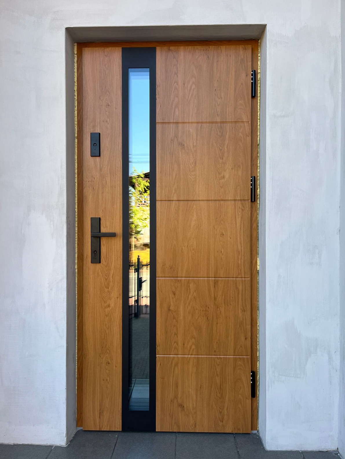 Nowoczesne drzwi wejściowe z drewnopodobnym panelem i pionowym przeszkleniem, czarne okucia, osadzone w surowym, jasnym murze.