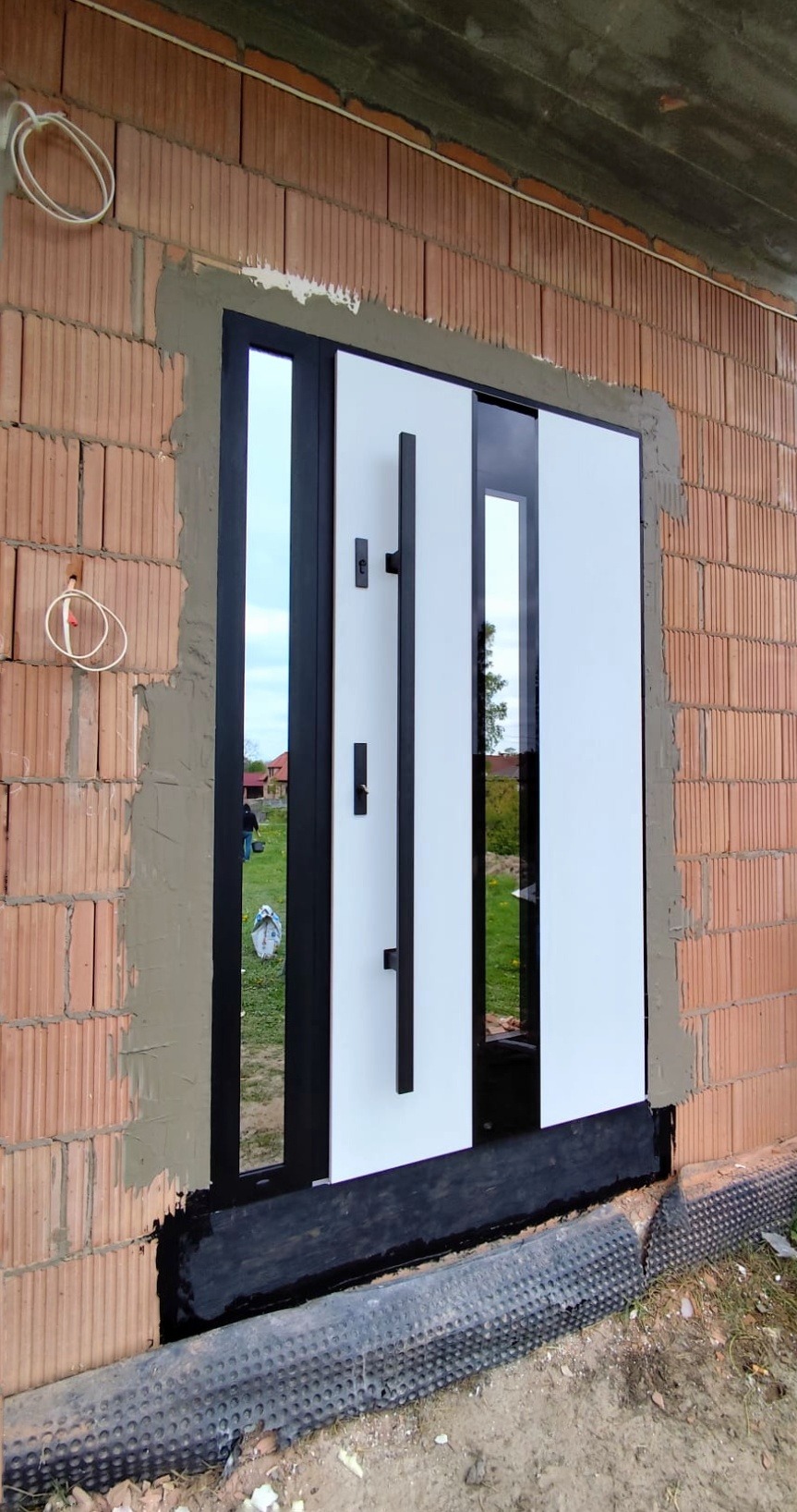 Montaż nowoczesnych drzwi przesuwnych z czarną ramą i białymi panelami w surowym murze z cegły. Widoczne detale montażu i otoczenie budowy.