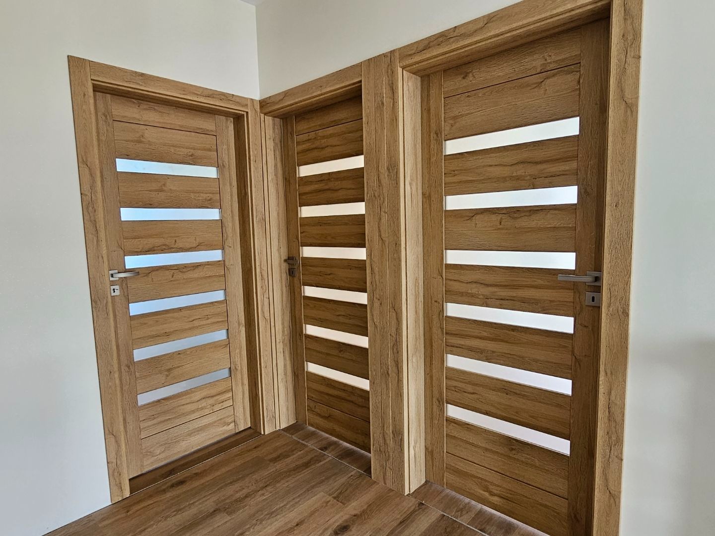 Trzy drewniane drzwi wewnętrzne z poziomymi przeszkleniami, zamontowane w jasnym pomieszczeniu z podłogą imitującą drewno. Widok z bliska, prezentujący detale wykończenia.