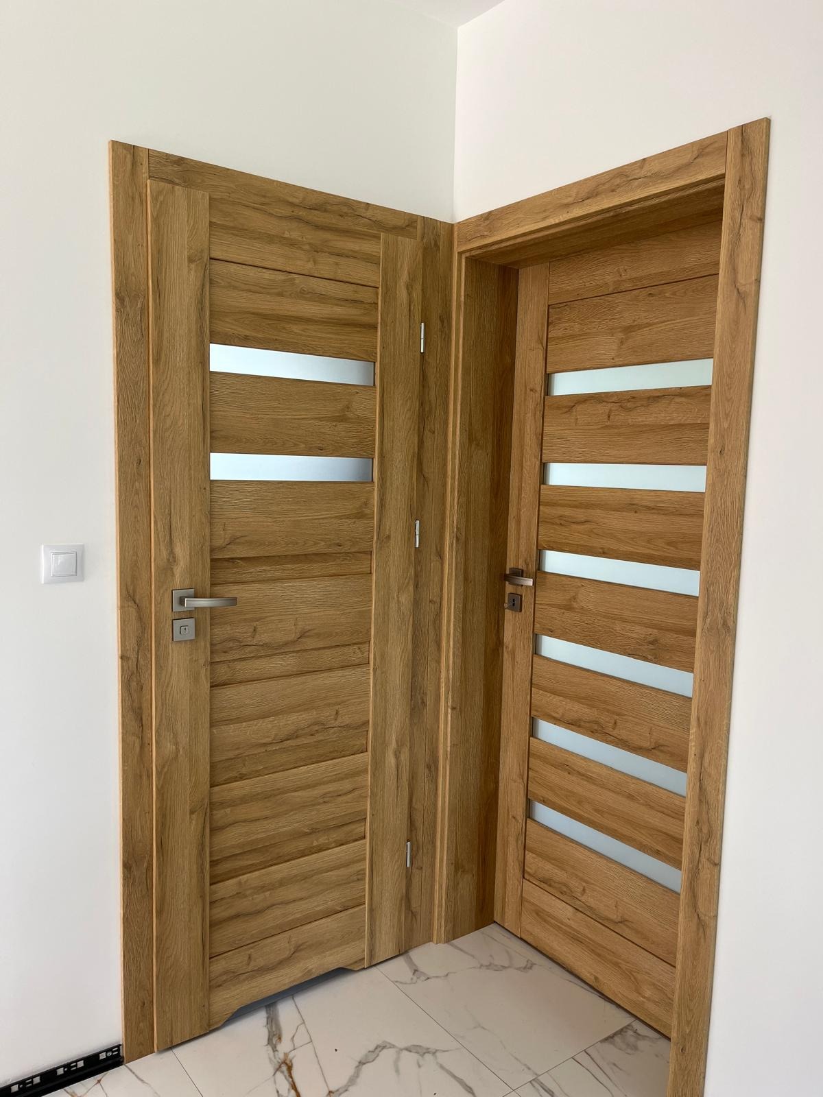Dwa drewniane skrzydła drzwiowe z poziomymi, matowymi przeszkleniami, zamontowane w białym wnętrzu. Widoczny fragment podłogi z marmurowym wzorem.