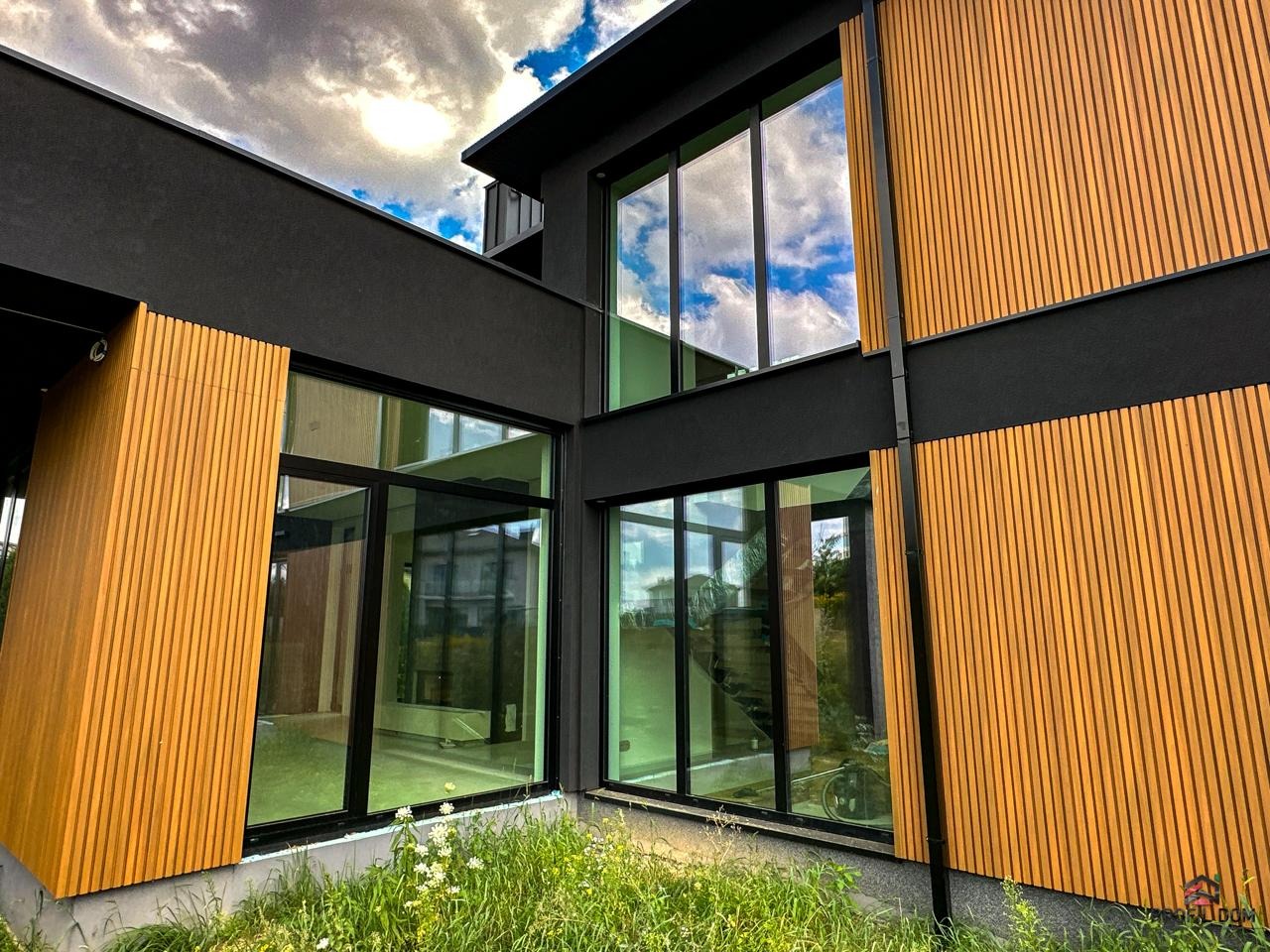 Nowoczesny dom z dużymi przeszkleniami i drewnianą elewacją w pionowe lamele. W oknach odbija się błękitne niebo z chmurami. Minimalistyczny design.