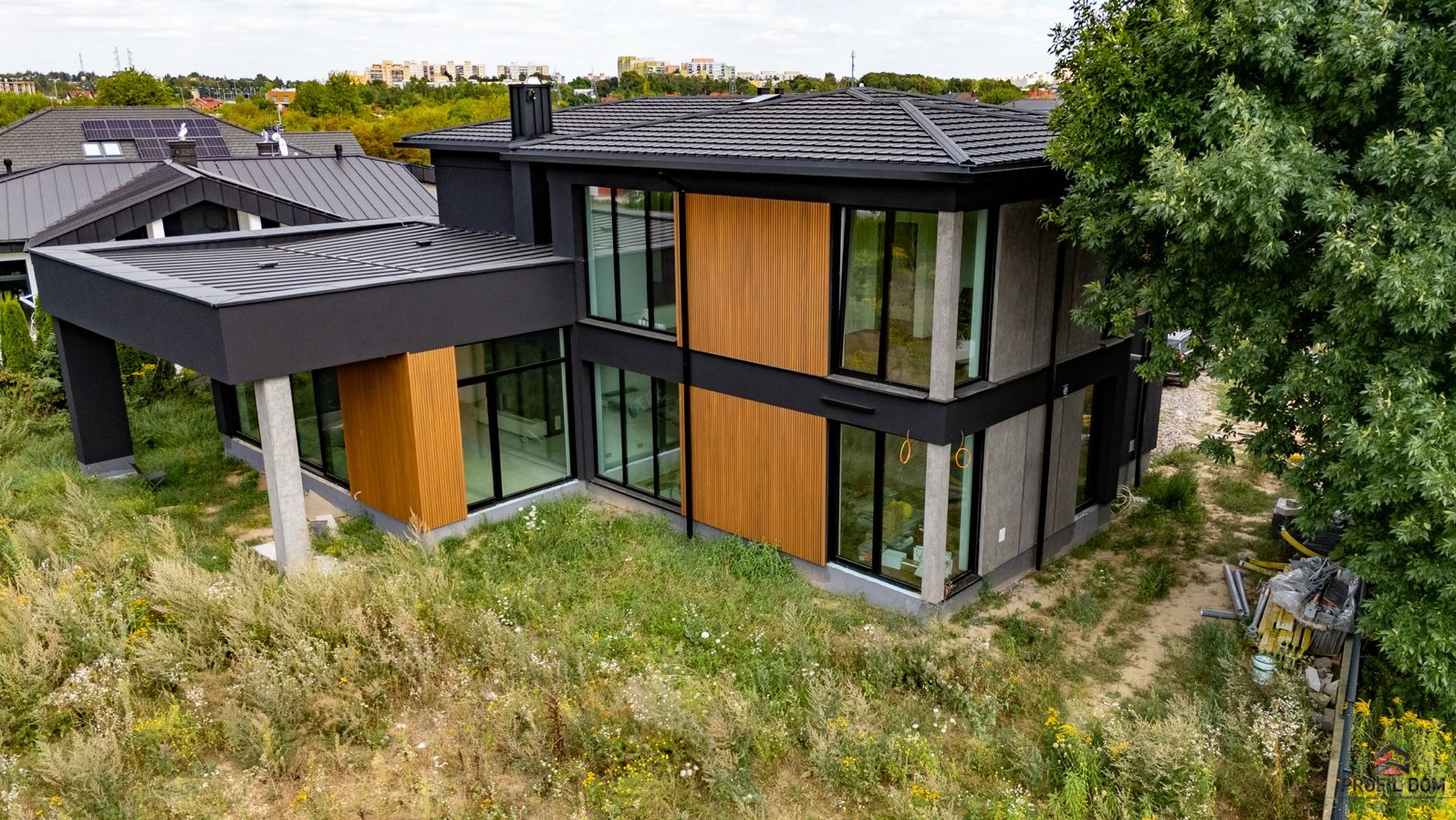 Nowoczesny, dwukondygnacyjny dom z dużymi przeszkleniami, wykończony drewnianą okładziną i szarą elewacją. Widok z góry na bujną roślinność wokół budynku.