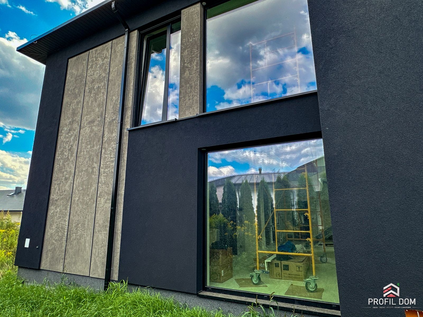 Nowoczesny dom z dużymi oknami, w których odbija się błękitne niebo. Elewacja w odcieniach szarości, z widocznym rusztowaniem wewnątrz, sugerującym prace wykończeniowe.