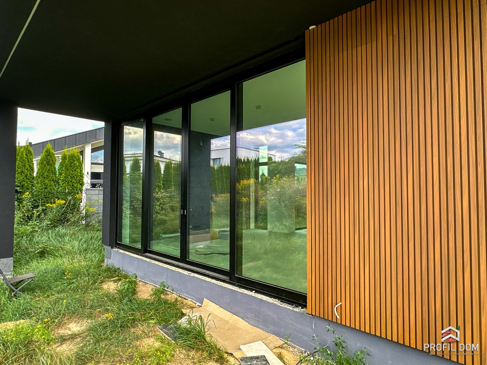 Nowoczesne, czarne okna przesuwne z widokiem na zieleń, połączone z drewnianą elewacją wertykalną. Minimalistyczny design, naturalne światło, trawa i krzewy w tle.