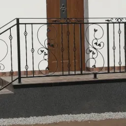 Czarna, metalowa balustrada schodowa z dekoracyjnymi elementami roślinnymi, prowadząca do drewnianych drzwi wejściowych w jasnym budynku.