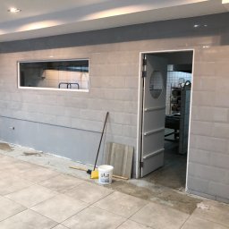 Stefbud - Wnętrze w trakcie prac wykończeniowych: szare płytki na ścianach, częściowo pomalowana dolna część ściany, widoczny otwór okienny do zaplecza, rozłożone płytki podłogowe, wiadro z farbą, szczotka...