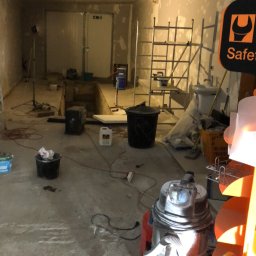 Stefbud - Piwnica w trakcie remontu, widoczne rusztowania, wiadro, pojemnik z gruntem, odkurzacz przemysłowy, ściany przygotowane do malowania, sufit z widocznymi nierównościami.