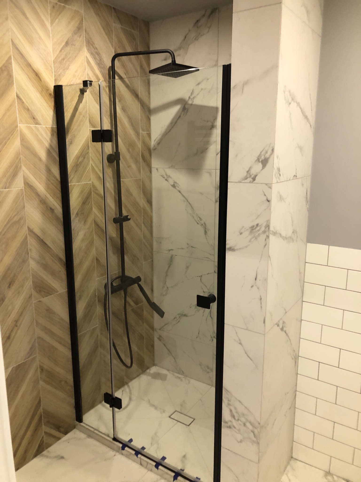 Nowoczesna kabina prysznicowa z czarnymi profilami i deszczownicą, wykończona płytkami imitującymi drewno ułożonymi w jodełkę oraz białym marmurem.