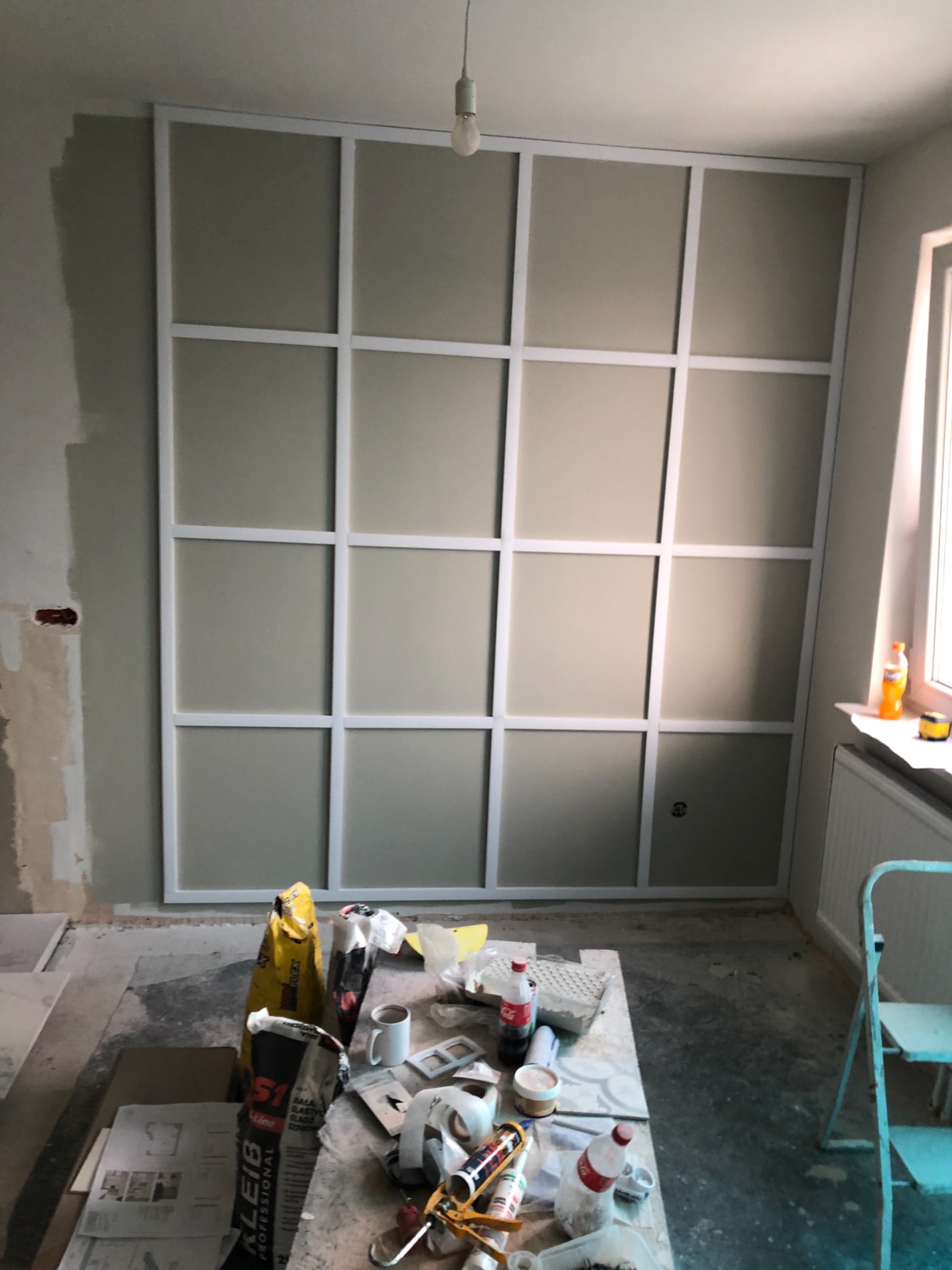 Ściana w trakcie montażu dekoracyjnych paneli z białymi listwami tworzącymi geometryczny wzór, widoczne materiały budowlane i narzędzia na pierwszym planie.