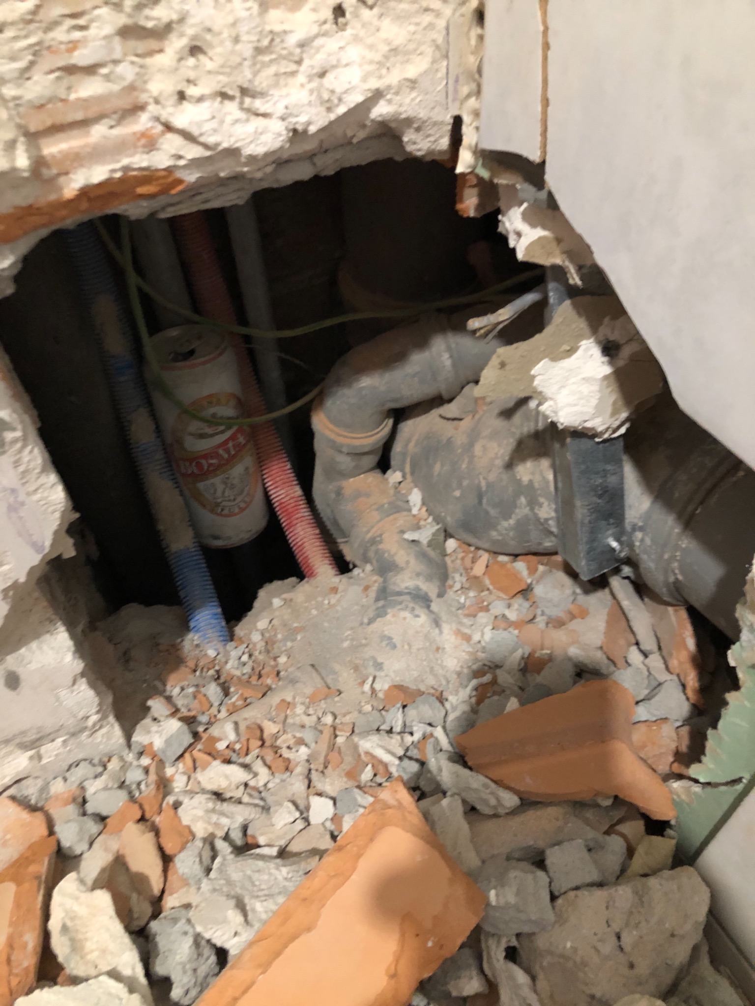 Otwór w ścianie z widocznymi rurami kanalizacyjnymi, przewodami i puszką po napoju wśród gruzu.