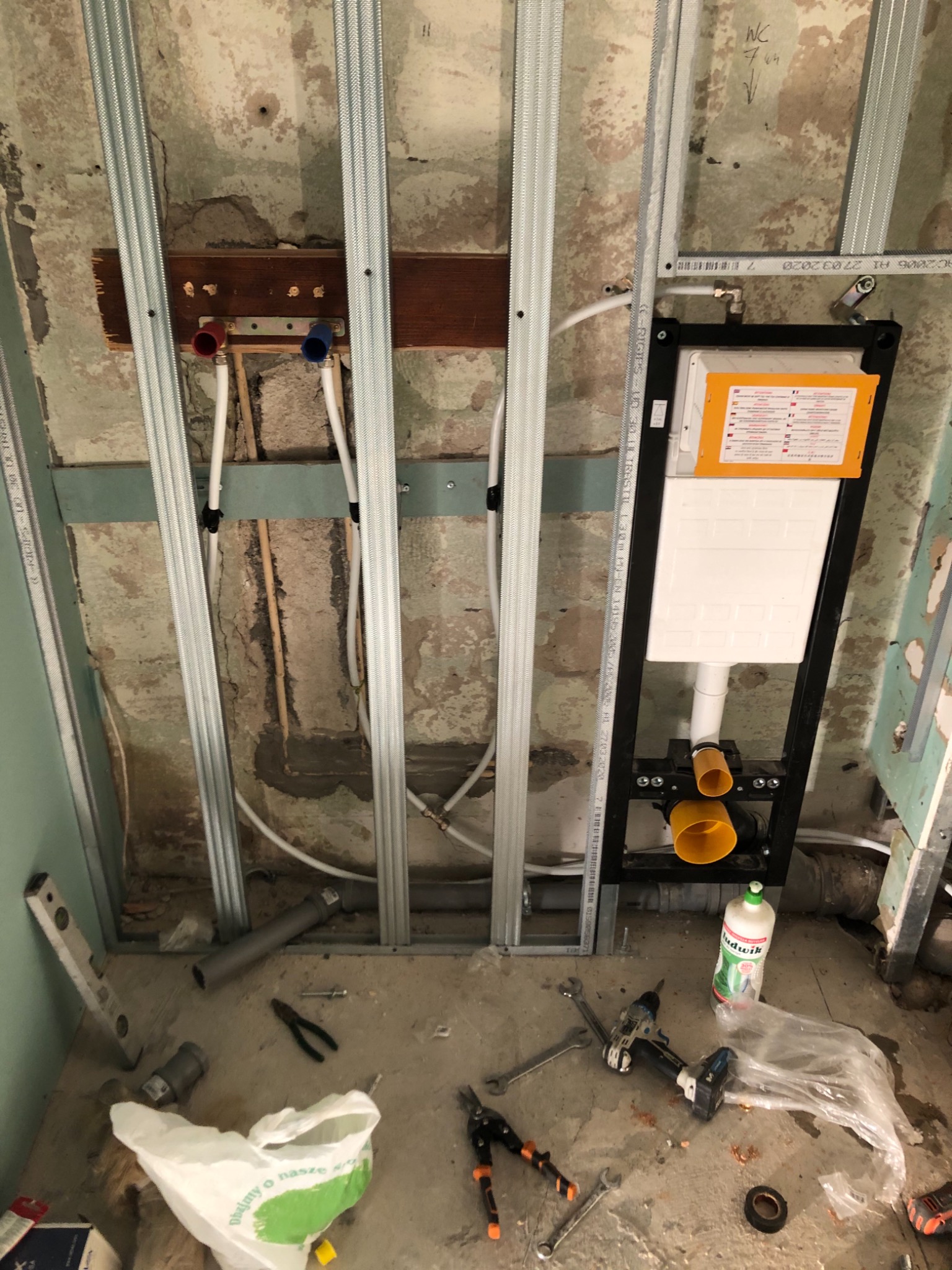 Montaż stelaża podtynkowego WC na tle surowej ściany z widocznymi rurami, elementami konstrukcyjnymi z profili metalowych oraz narzędziami i preparatem czyszczącym Ludwik na podłodze.