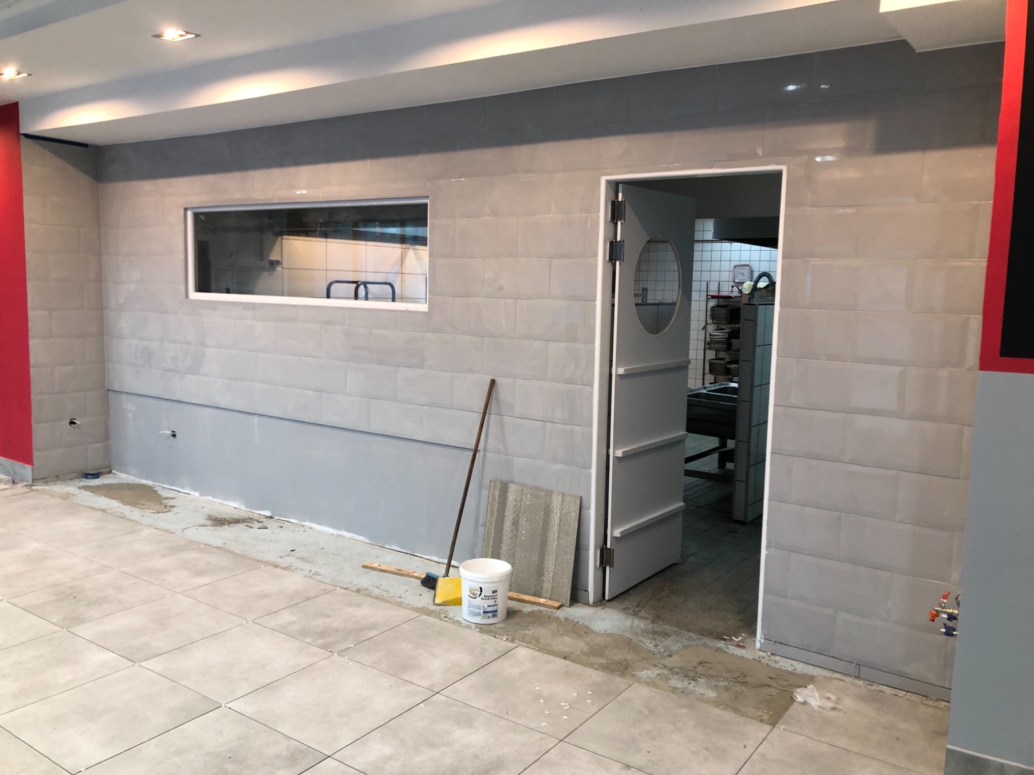 Wnętrze w trakcie prac wykończeniowych: szare płytki na ścianach, częściowo pomalowana dolna część ściany, widoczny otwór okienny do zaplecza, rozłożone płytki podłogowe, wiadro z farbą, szczotka...