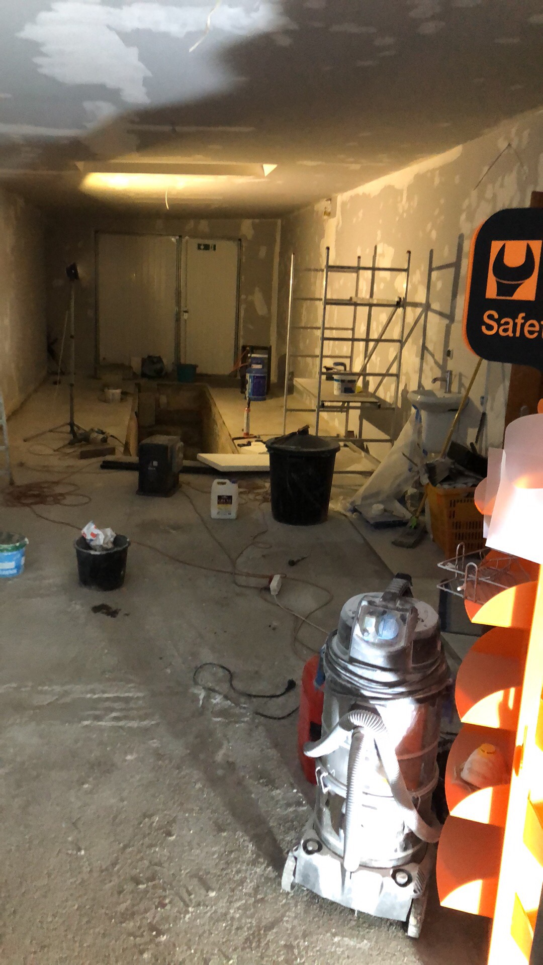 Piwnica w trakcie remontu, widoczne rusztowania, wiadro, pojemnik z gruntem, odkurzacz przemysłowy, ściany przygotowane do malowania, sufit z widocznymi nierównościami.