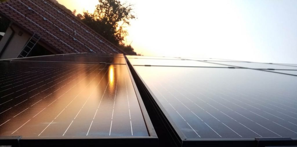 Błyszczące panele słoneczne na dachu domu, odbijające zachodzące słońce, z widoczną drabiną opartą o ścianę.