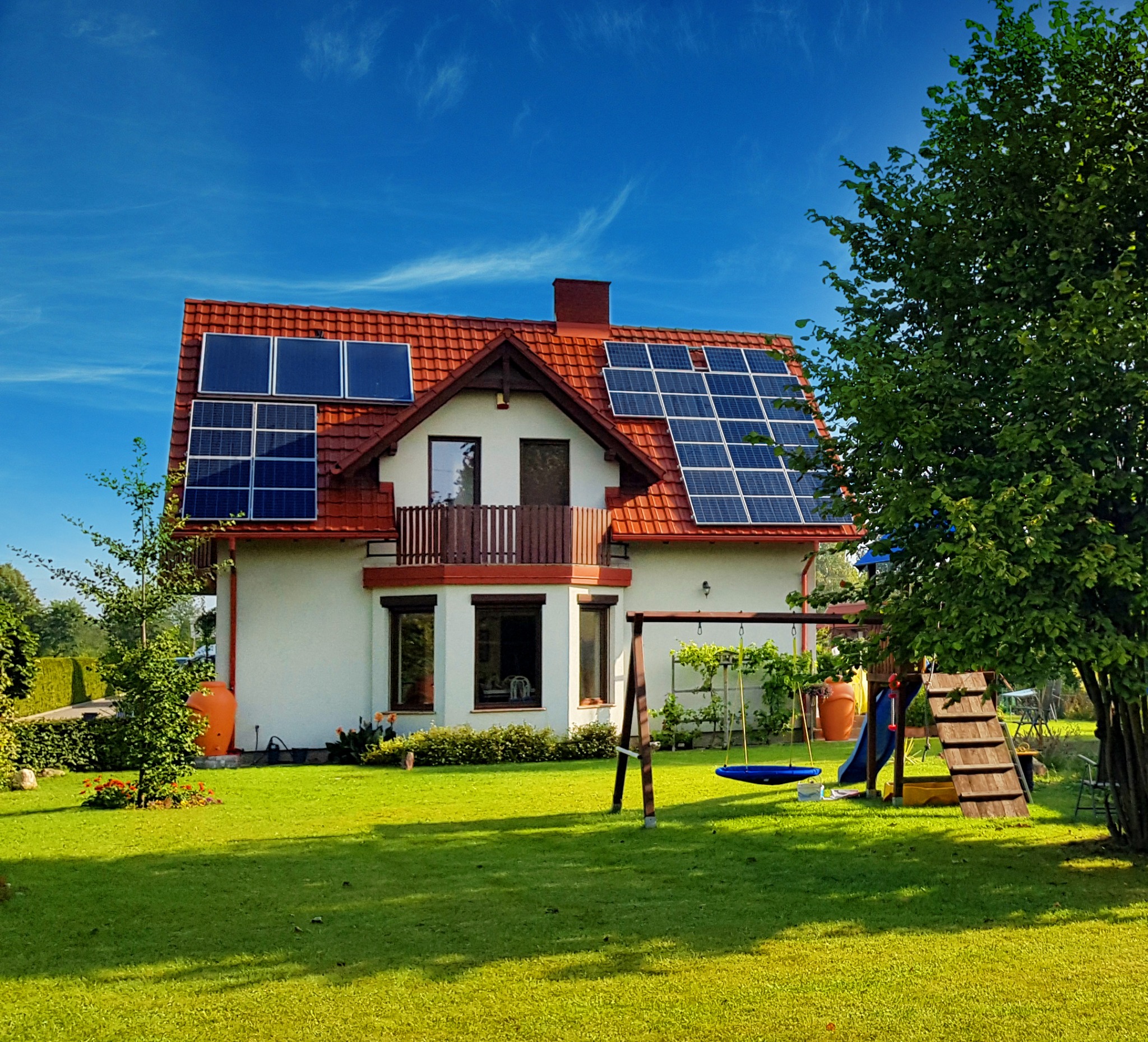 Dom jednorodzinny z czerwonym dachem i panelami słonecznymi na dachu, otoczony zielonym trawnikiem i drzewami, błękitne niebo w tle.