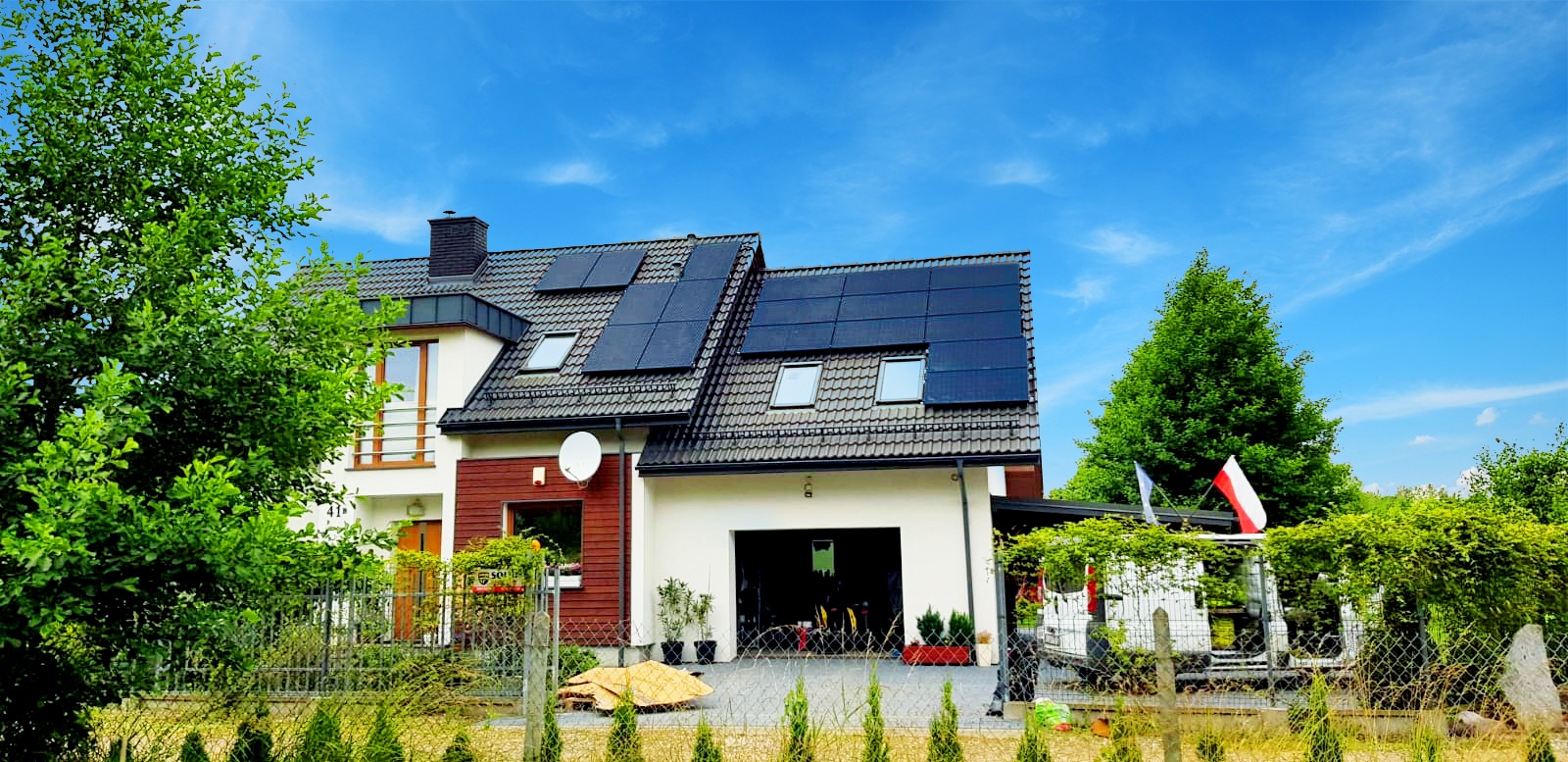 Dwukondygnacyjny dom jednorodzinny z panelami słonecznymi na dachu, widoczny zza ogrodzenia i zieleni, z flagą Polski powiewającą w tle.