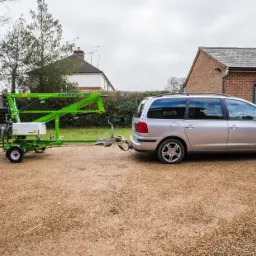 Zielony podnośnik koszowy Nifty 90 podpięty do srebrnego samochodu osobowego na żwirowym podjeździe przed domem z cegły.