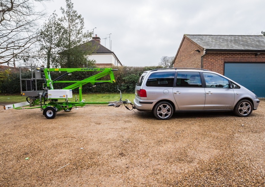 Zielony podnośnik koszowy Nifty 90 podpięty do srebrnego samochodu osobowego na żwirowym podjeździe przed domem z cegły.