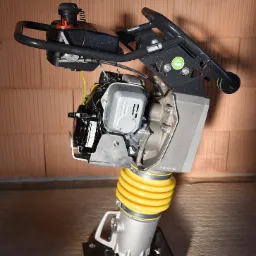 Stacjonarna ubijarka stopowa z silnikiem spalinowym i żółtą osłoną amortyzującą, ustawiona na betonowej powierzchni, na tle ściany z bloczków ceramicznych.