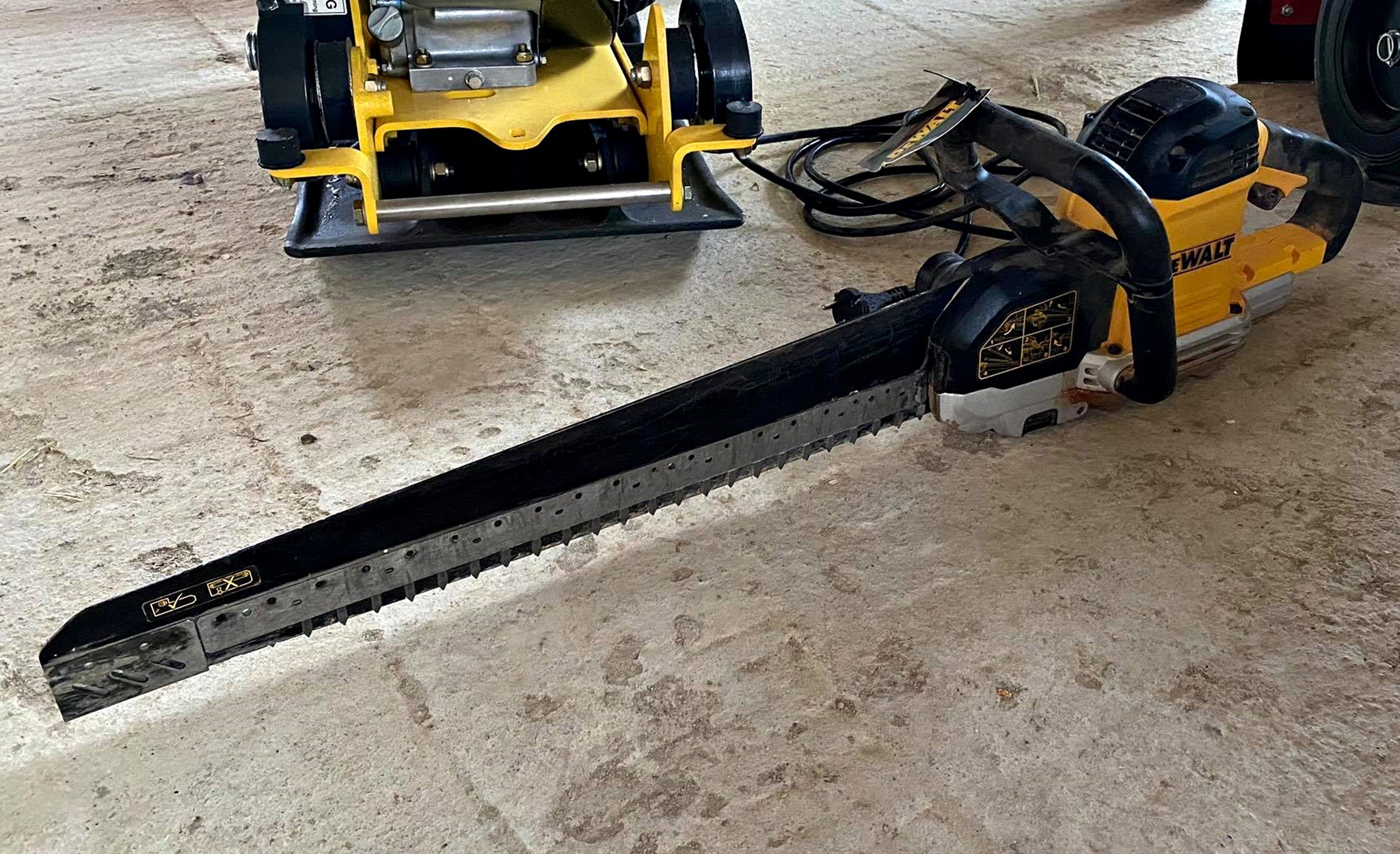 Elektryczna piła łańcuchowa DeWalt leżąca na betonowej posadzce obok żółtej zagęszczarki wibracyjnej