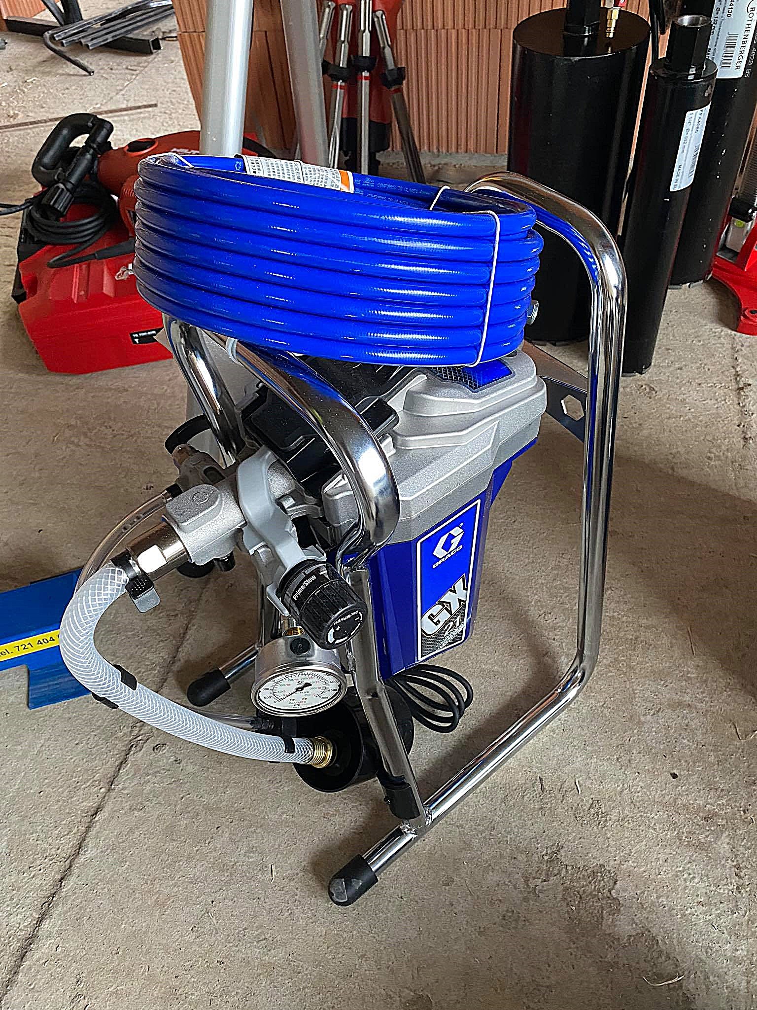 Agregat malarski Graco GX 21 z nawiniętym niebieskim wężem, widoczny manometr i elementy sterujące, w tle wiertnica i akcesoria budowlane.