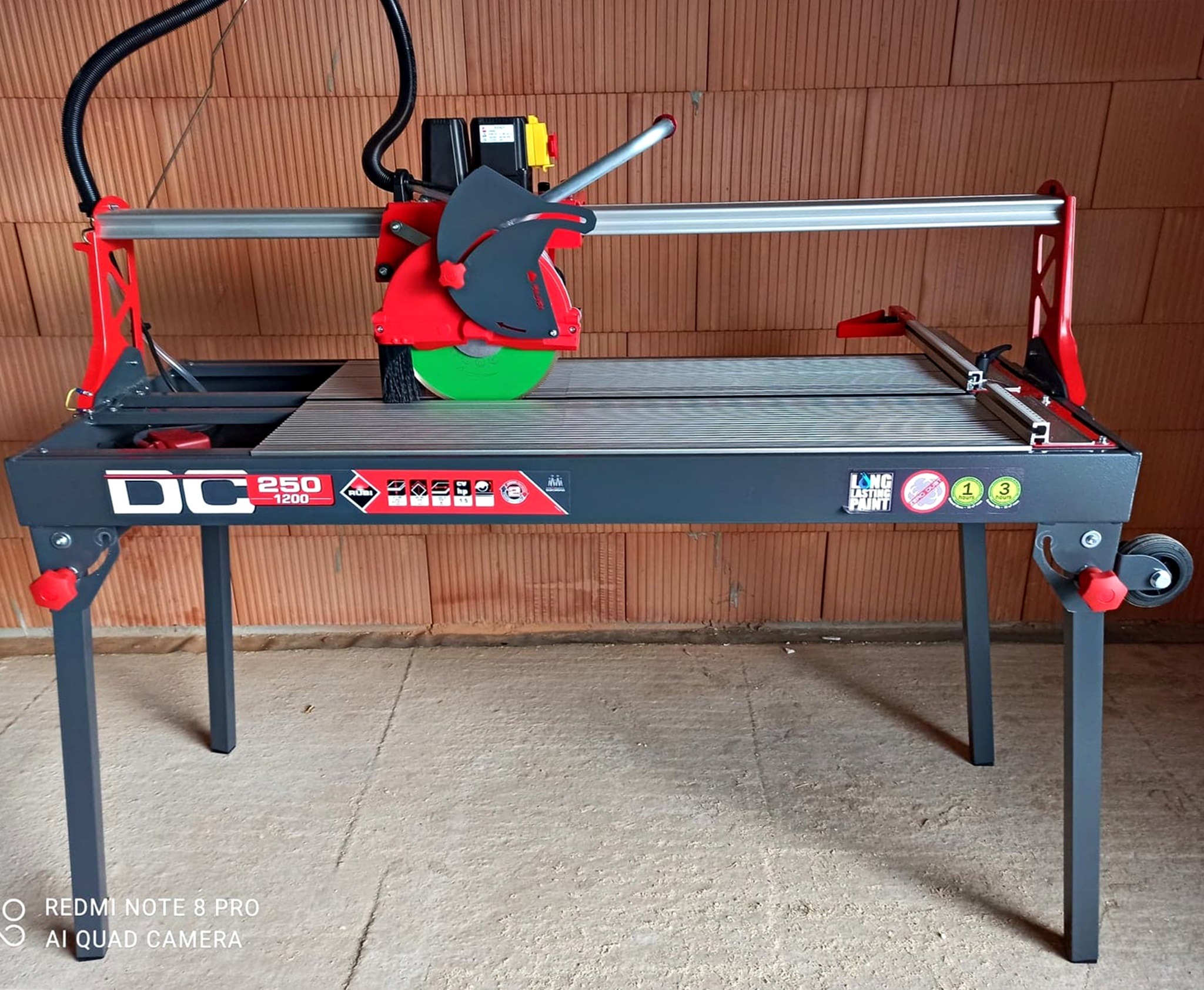 Profesjonalna przecinarka do płytek Rubí DC-250 1200 z tarczą diamentową, ustawiona na posadzce, z tłem z cegieł dziurawki.