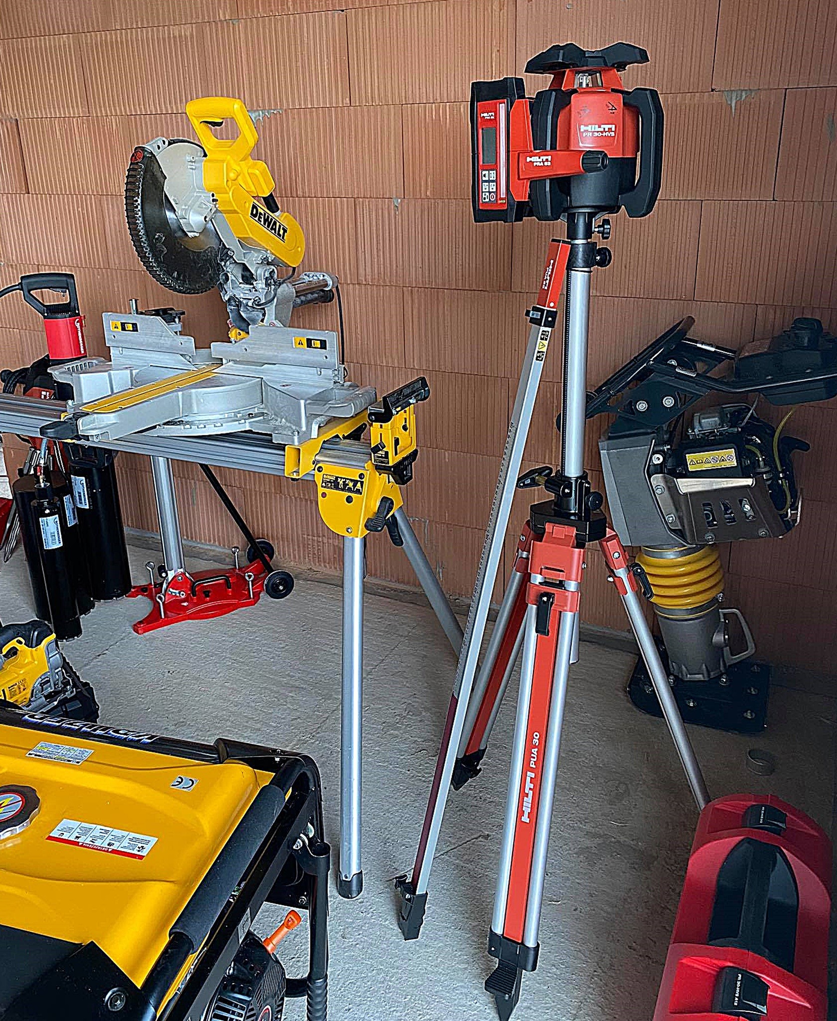 Zestaw narzędzi budowlanych: piła ukośnica DeWalt na stole roboczym, niwelator laserowy Hilti na statywie, ubijak wibracyjny oraz agregat prądotwórczy, na tle ściany z cegły.