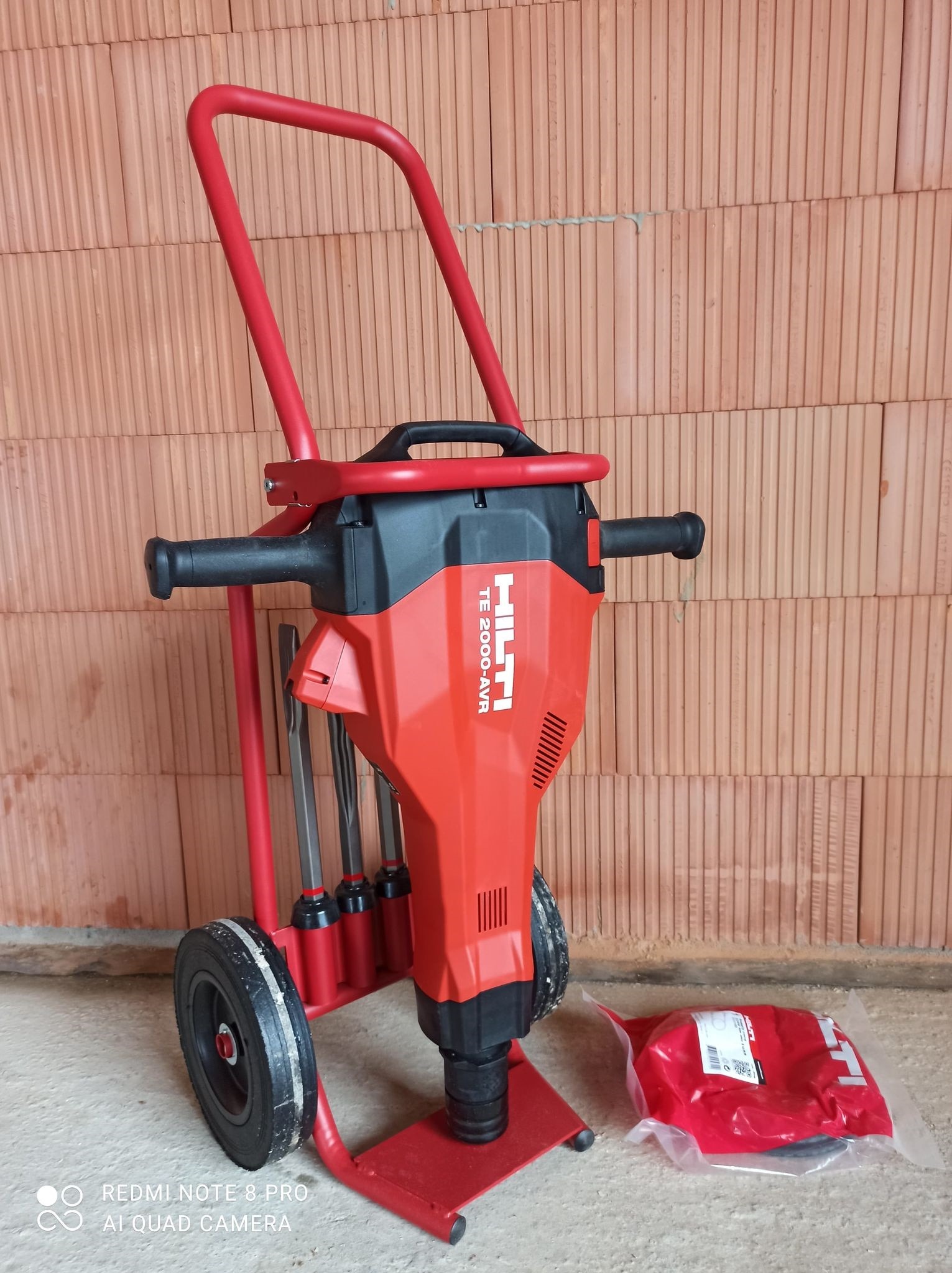 Czerwony młot wyburzeniowy Hilti TE 2000-AVR na wózku transportowym, z dodatkowymi dłutami, na tle ściany z cegły dziurawki.