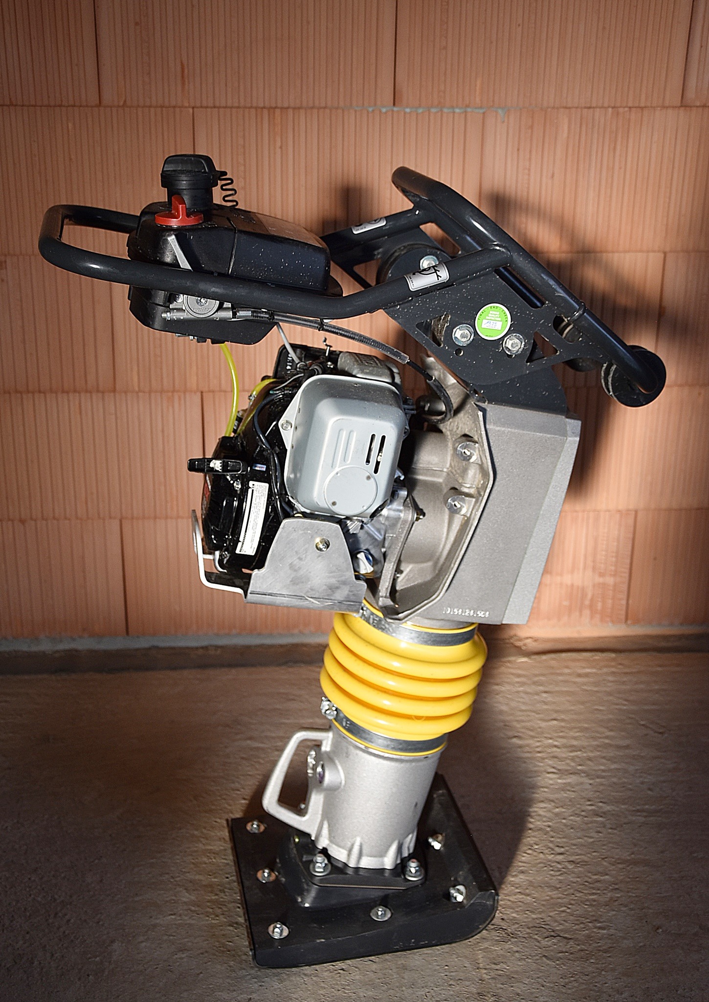 Stacjonarna ubijarka stopowa z silnikiem spalinowym i żółtą osłoną amortyzującą, ustawiona na betonowej powierzchni, na tle ściany z bloczków ceramicznych.