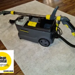 Profesjonalny odkurzacz piorący Kärcher Puzzi 10/1 na białym dywanie, widoczne logo Clean Rental w lewym dolnym rogu.