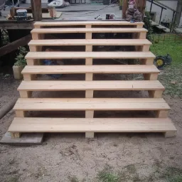 Surowe drewniane schody prowadzące z ziemi na podwyższony taras, widoczne elementy konstrukcyjne i otoczenie w trakcie budowy.