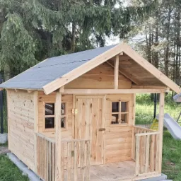 Nowa, surowa drewniana altana dla dzieci z małym gankiem i szarym dachem bitumicznym, umieszczona na trawie z widoczną zjeżdżalnią w tle.