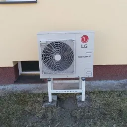 Biała zewnętrzna jednostka pompy ciepła LG Therma V zamontowana na metalowym stelażu przy budynku z beżową elewacją i małym oknem piwnicznym.