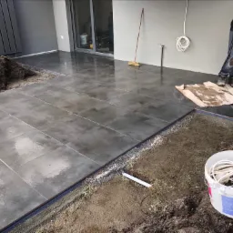 Układanie szarych płytek na zewnątrz budynku, widoczny fragment przygotowanego podłoża z drobnym żwirem, obok leży biała rura PCV, w tle stos ziemi i osoba w roboczych ubraniach.