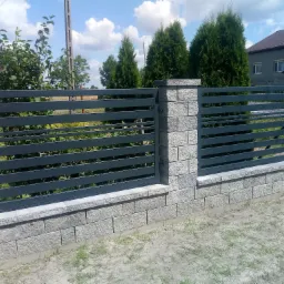 Nowoczesne, poziome, szare ogrodzenie panelowe z elementami murowanymi z szarej kostki, widoczne drzewa i dom w tle, słoneczny dzień.