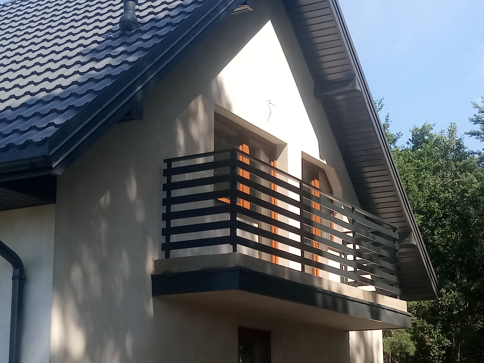 Czarna, pozioma balustrada balkonowa na tle jasnej elewacji domu z ciemnym dachem, widziana z dołu pod kątem.