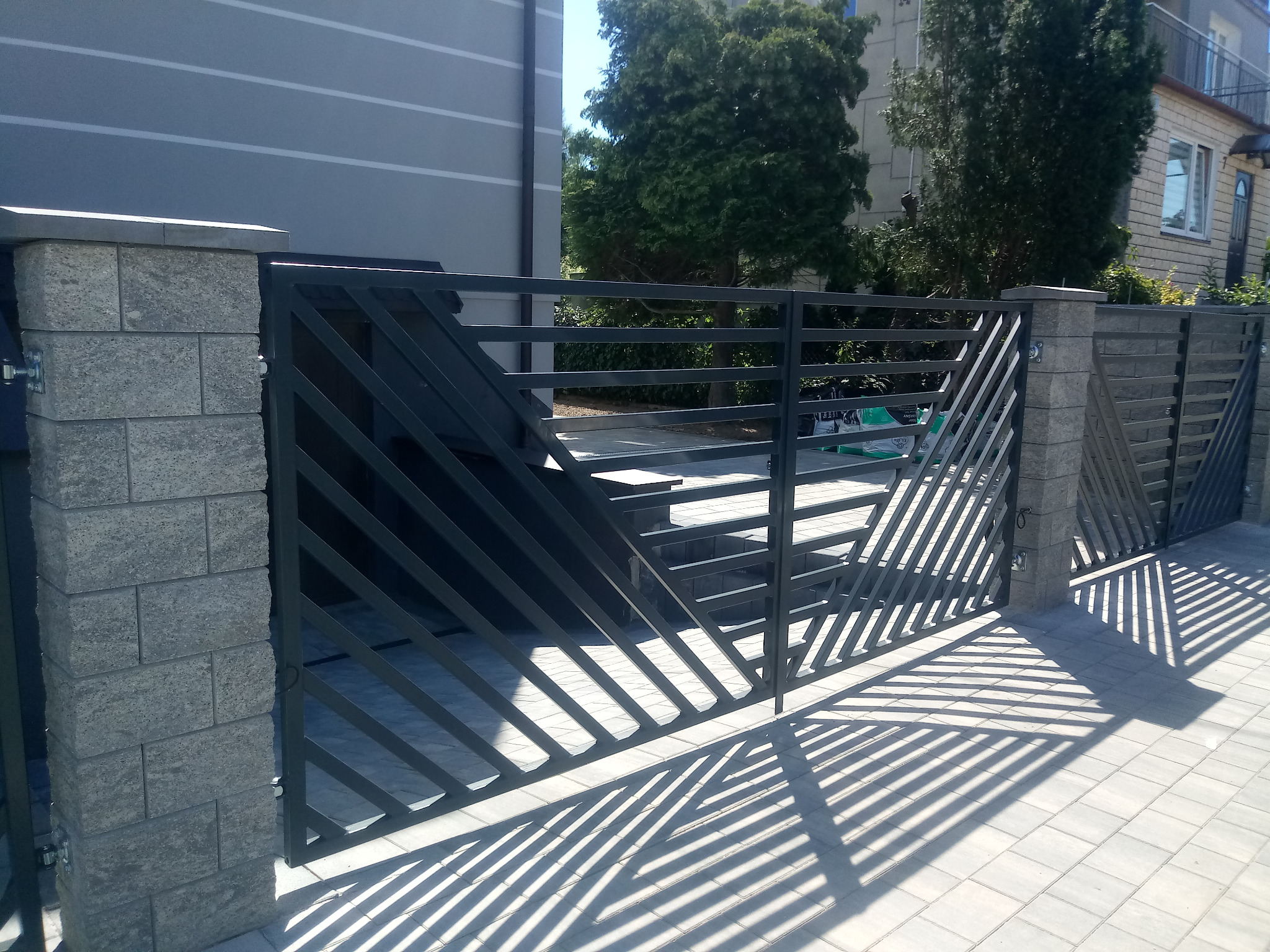 Nowoczesna brama wjazdowa z szarego metalu z ukośnymi szczeblami, osadzona na słupkach z szarej kostki, rzucająca geometryczny cień na jasną kostkę brukową.