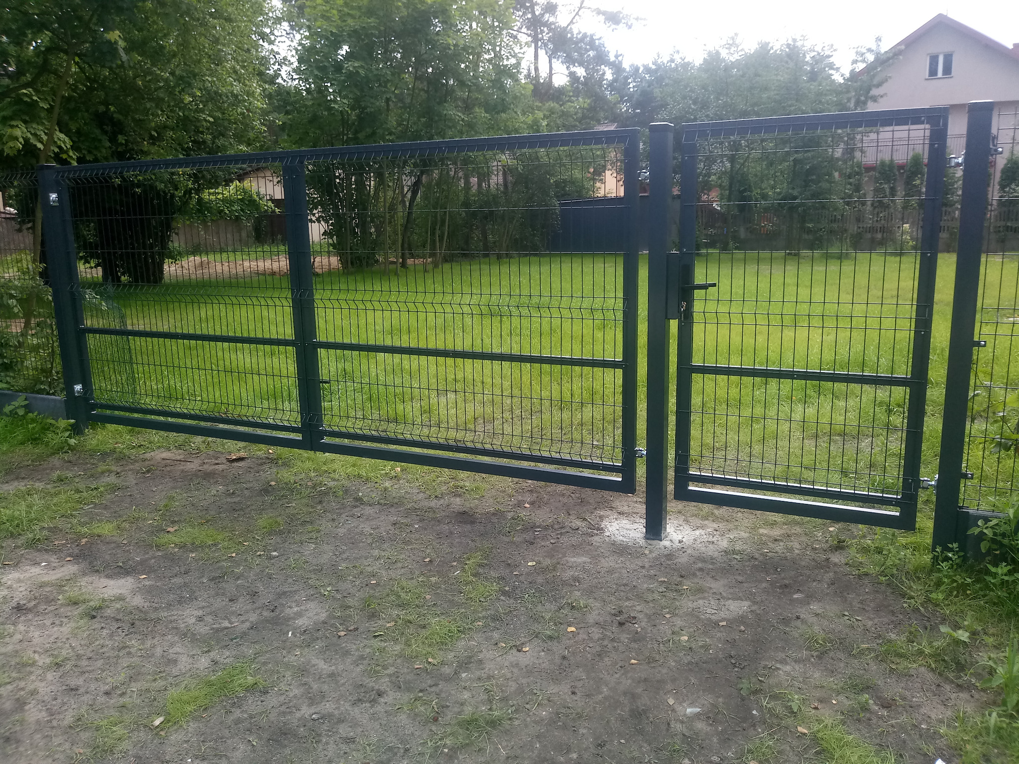 Nowoczesna, metalowa, panelowa brama w kolorze grafitowym, z widocznym zamkiem i klamką, otwierająca się na zadbany trawnik przed domem jednorodzinnym.