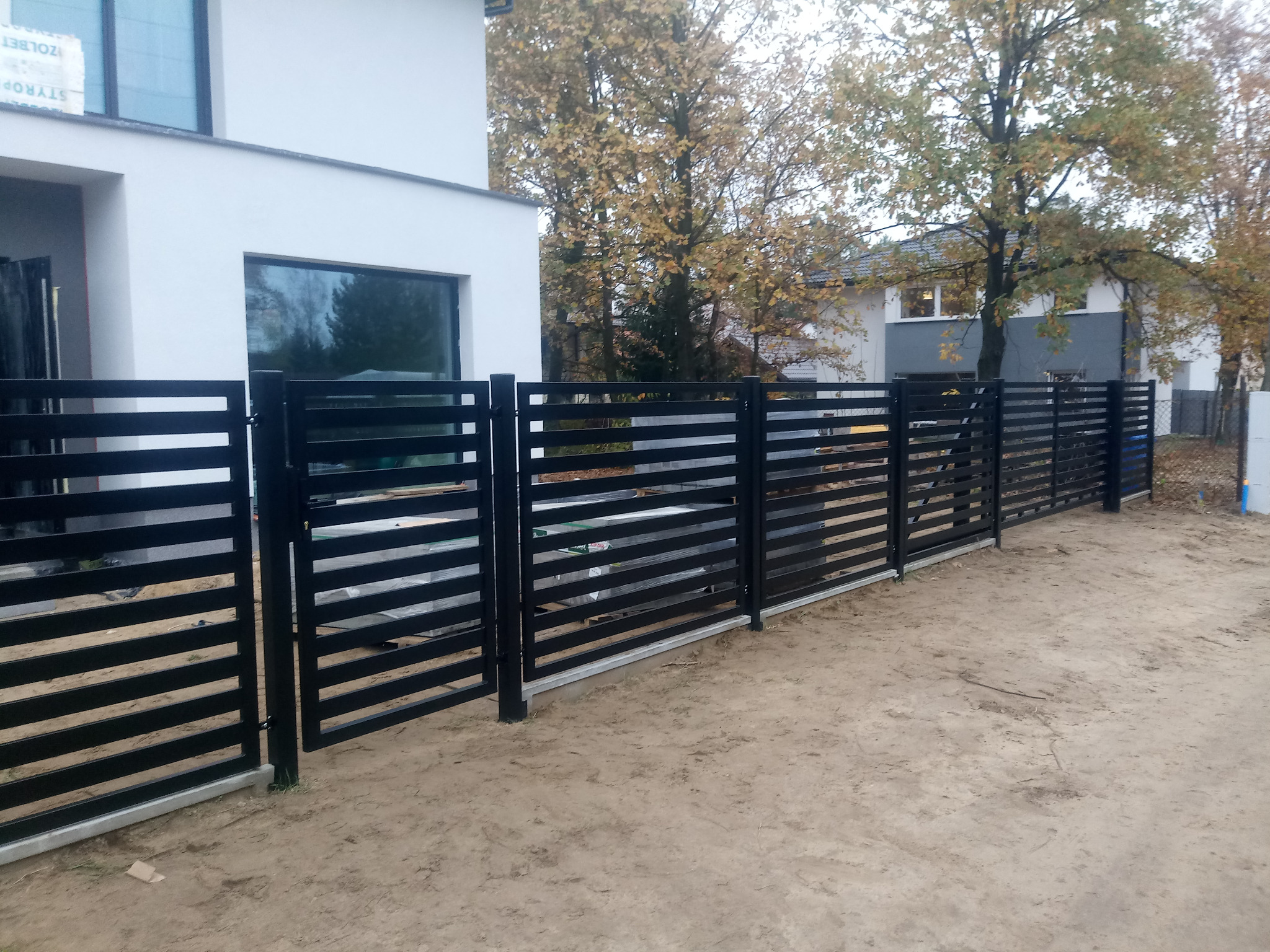 Nowoczesne, poziome, czarne ogrodzenie panelowe na betonowej podmurówce przed białym domem z dużym oknem, widok z zewnątrz posesji, jesienne drzewa w tle.