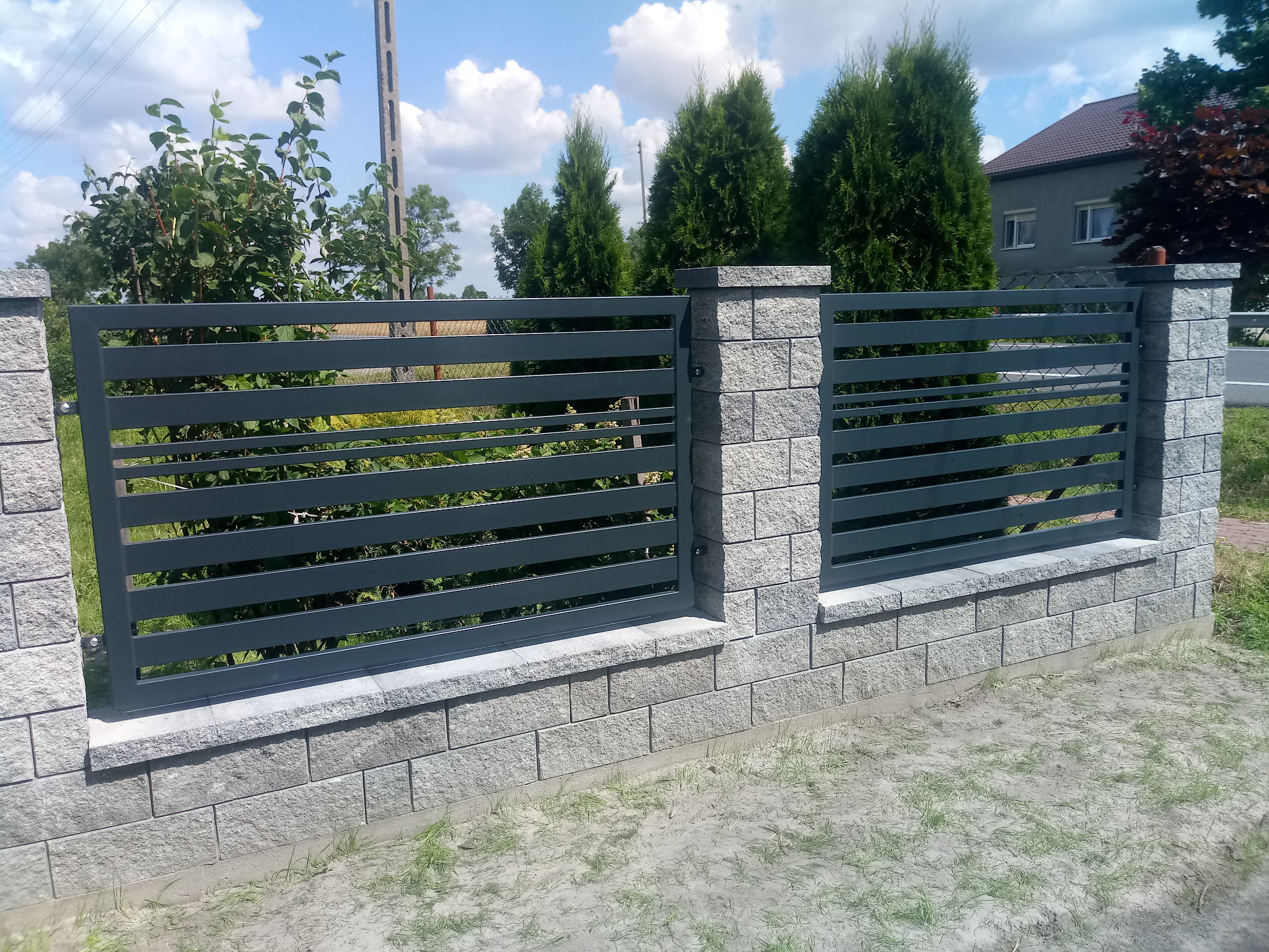 Nowoczesne, poziome, szare ogrodzenie panelowe z elementami murowanymi z szarej kostki, widoczne drzewa i dom w tle, słoneczny dzień.