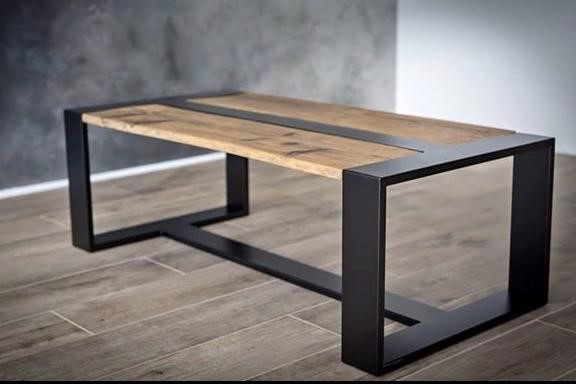 Stolik kawowy z blatem z jasnego drewna z ciemnym pasem pośrodku i czarną, metalową ramą o geometrycznym kształcie na tle szarej ściany i podłogi z płytek imitujących drewno.