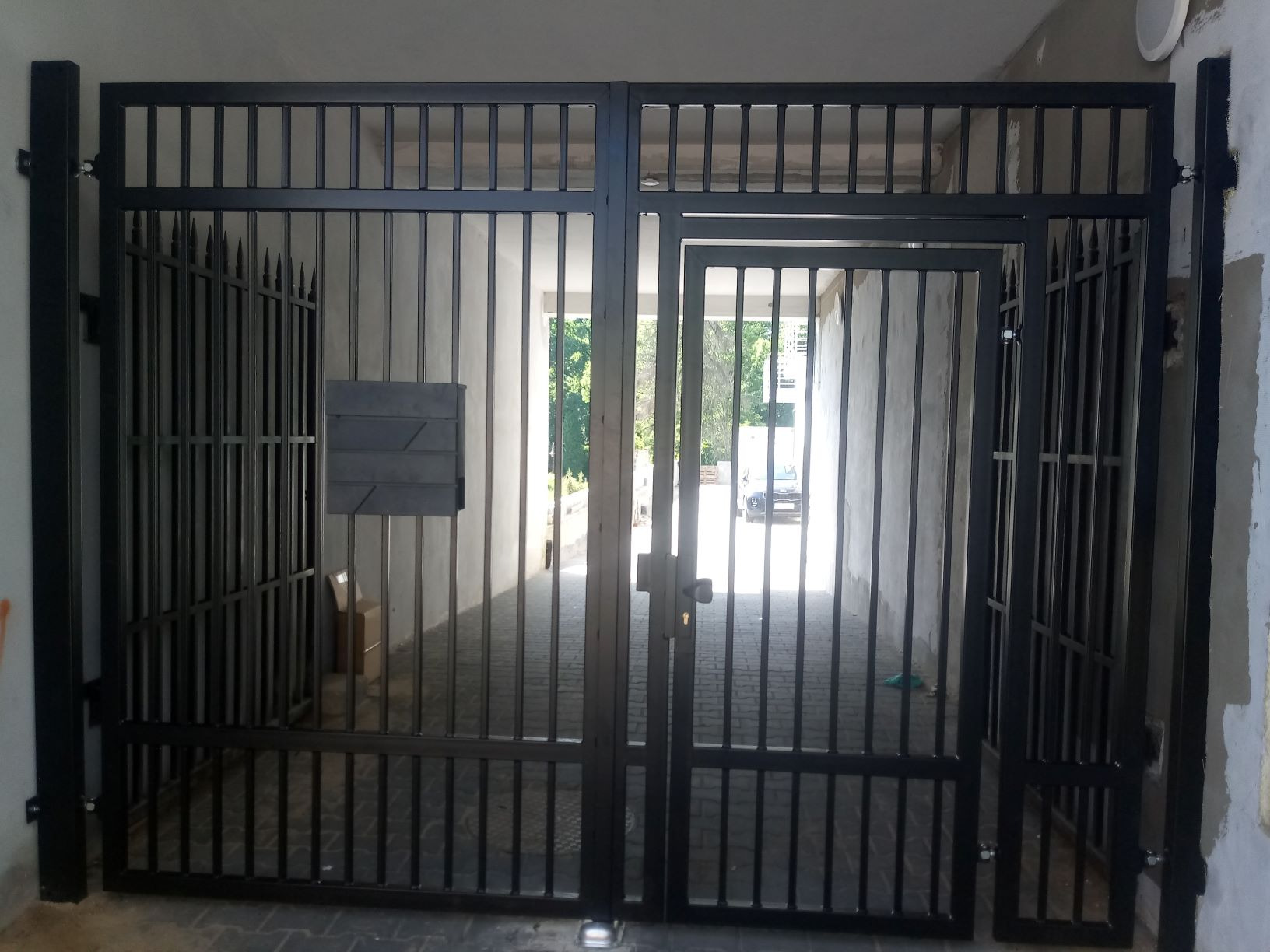 Czarna, metalowa brama dwuskrzydłowa z furtką, widok z zewnątrz na podwórze z kostką brukową i zaparkowanym samochodem w oddali.