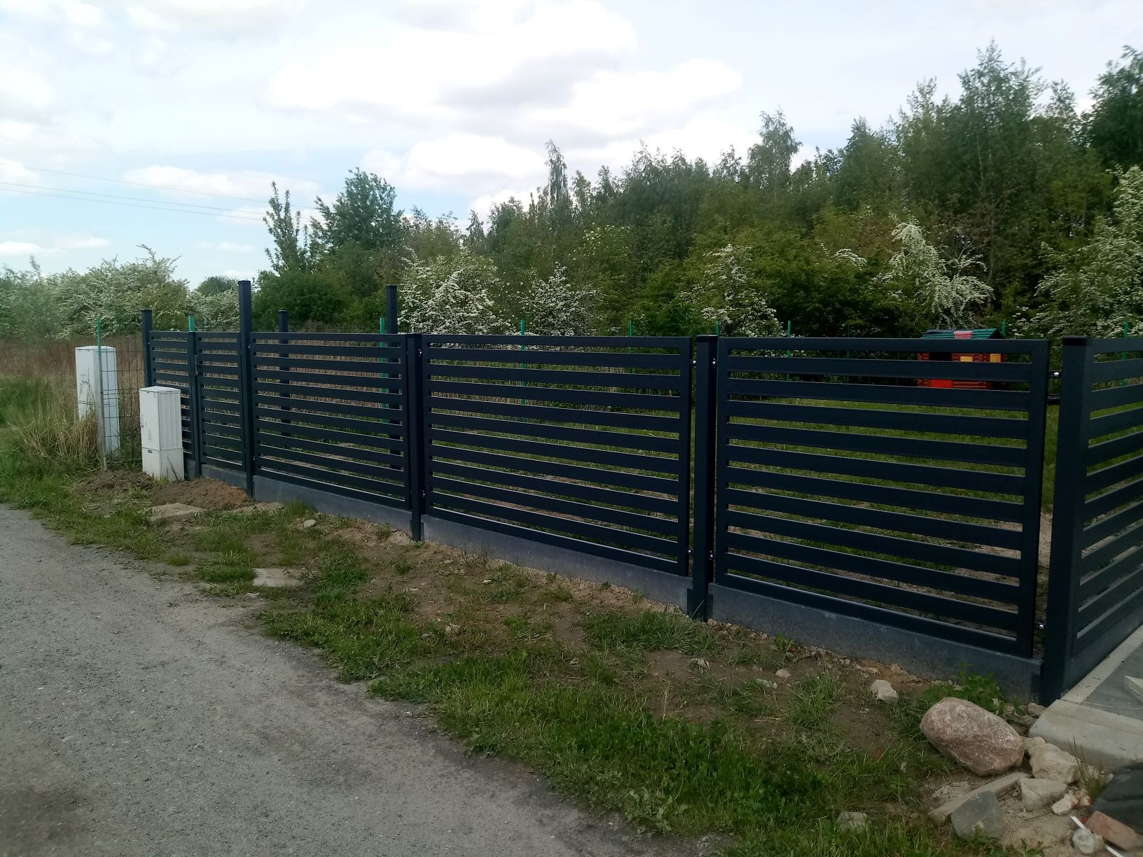 Nowoczesne, poziome ogrodzenie panelowe w kolorze grafitowym, z widocznymi słupkami i podmurówką, na tle zieleni i błękitnego nieba z chmurami.