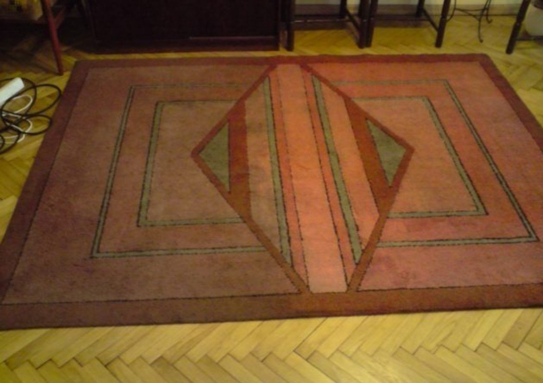 Prostokątny dywan w odcieniach czerwieni i brązu z geometrycznym wzorem, leżący na parkiecie ułożonym w jodełkę.